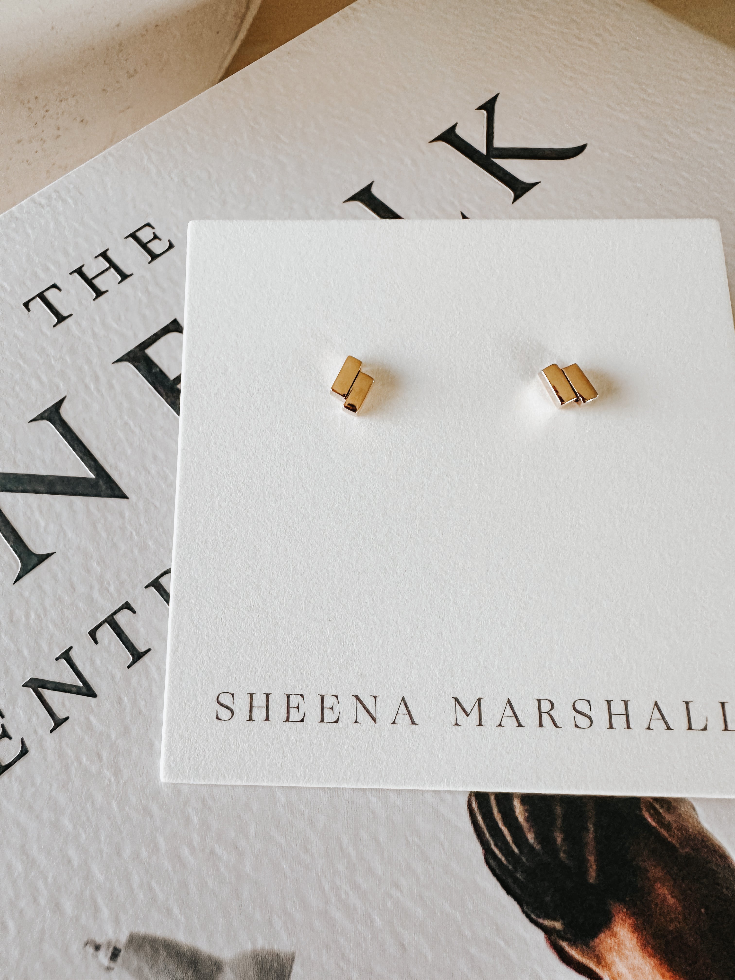 Close-up of solid gold Off-Balance Bar Stud Earrings with an asymmetrical design, symbolizing resilience and balance during life's uncertain moments, displayed on a Sheena Marshall jewelry card.