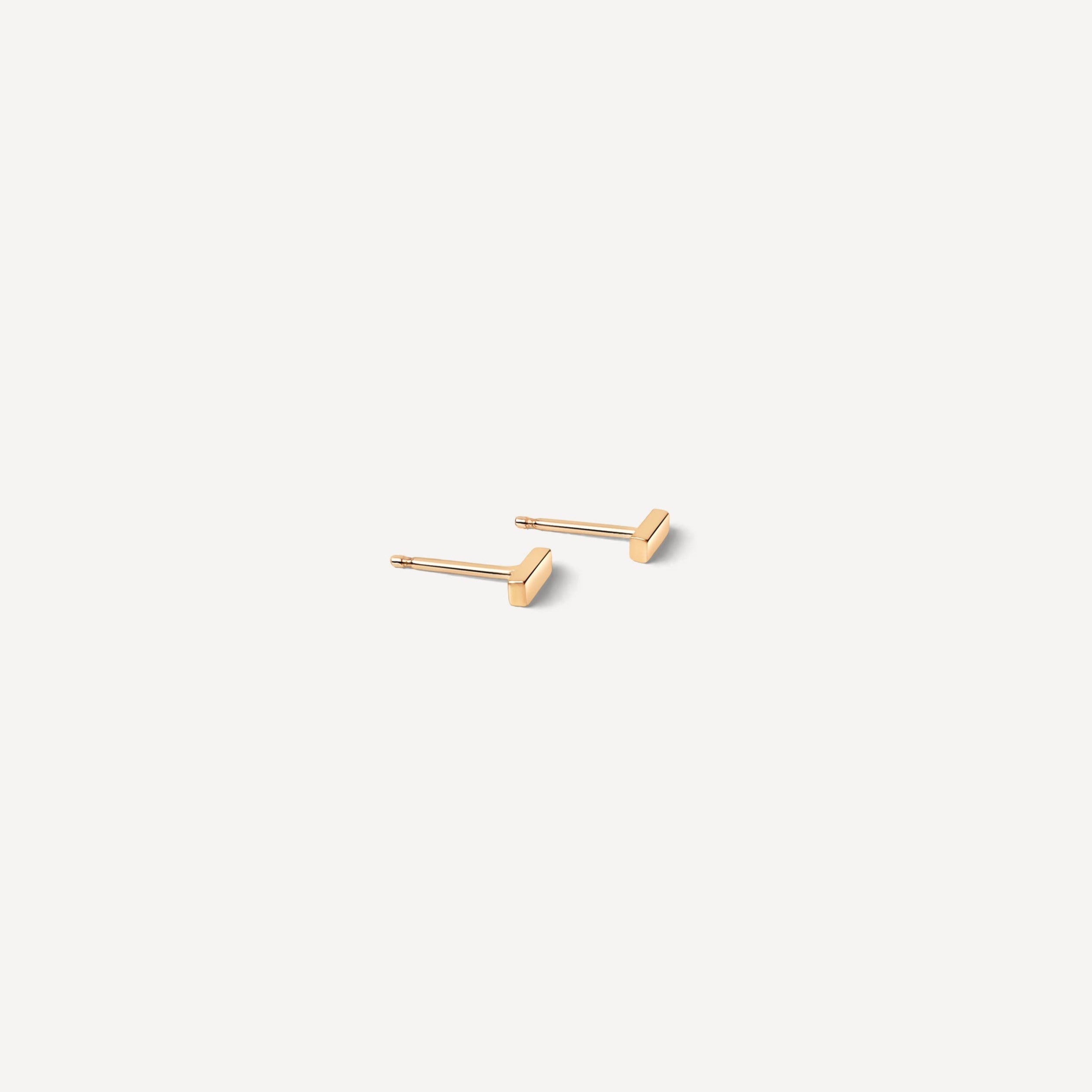 Flat lay with white background of simple gold bar studs shown from angle, highlighting delicate design for those seeking minimal gold jewelry pieces.