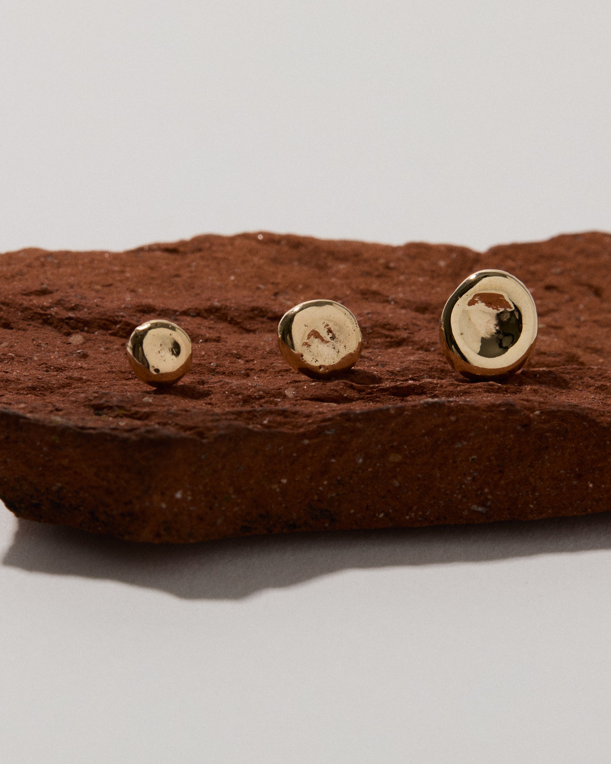 Three handcrafted 14k gold Molten Pebble studs with unique organic texture displayed on red clay stone, showing small, medium, and large size options of sculptural earrings