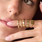 A close-up of a model's hand and face wearing a stack of five gold rings, including textured bands, a beaded ring, and a square face ring.