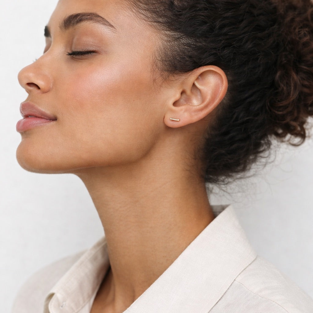 Woman with styled hair wearing a light-colored shirt against a plain background wearing simple gold fill bar stud earrings. 