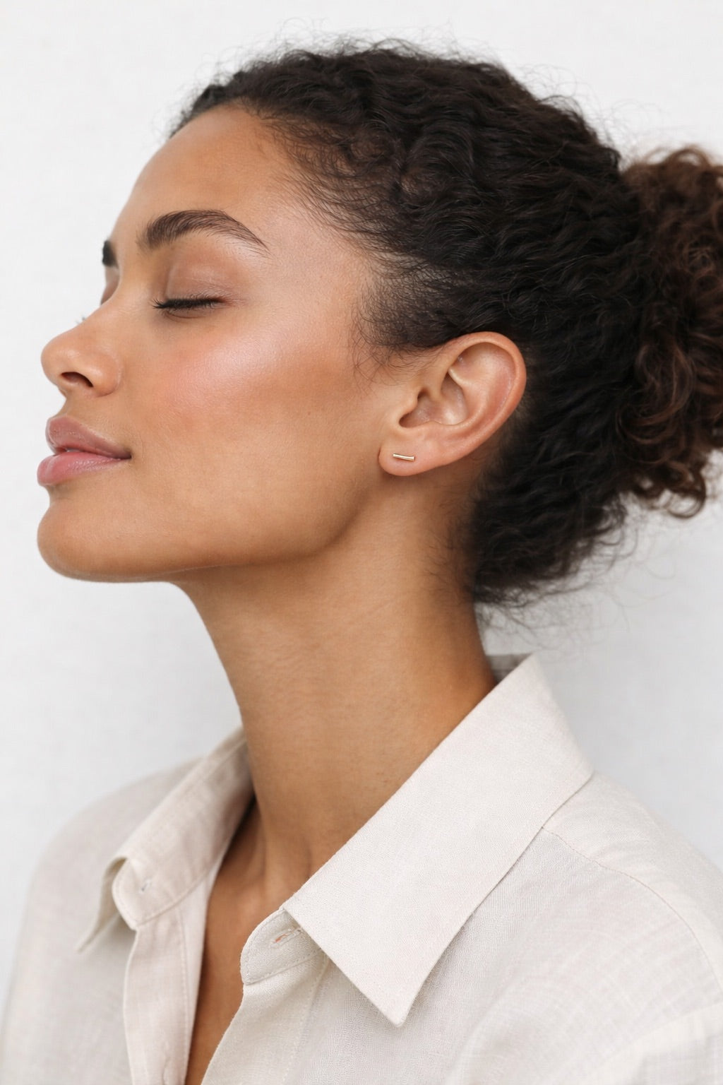 Woman with styled hair wearing a light-colored shirt against a plain background wearing simple gold fill bar stud earrings. 