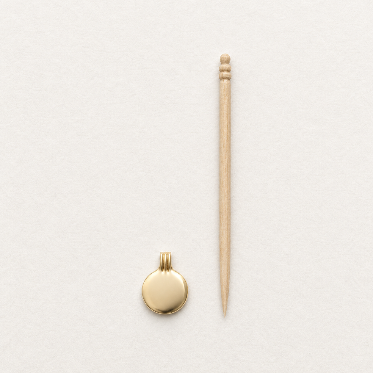 A small round gold pendant with a bail and a light wood toothpick placed vertically beside it on a clean white textured background.