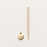 A small round gold pendant with a bail and a light wood toothpick placed vertically beside it on a clean white textured background.