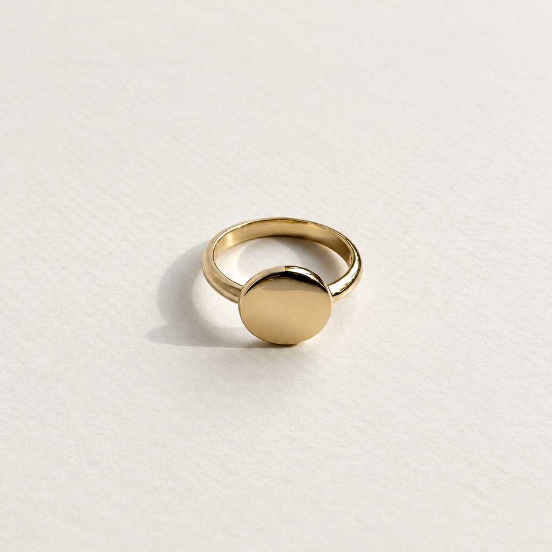 A minimalist gold signet ring with a flat circular disc, photographed on a clean, textured white surface.