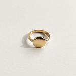 A minimalist gold signet ring with a flat circular disc, photographed on a clean, textured white surface.