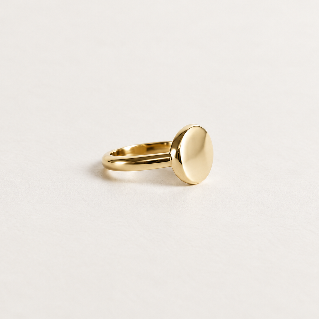A minimalist gold signet ring with a flat, oval disc face, photographed on a clean, light off-white background.
