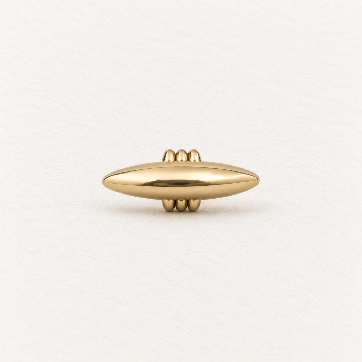 A polished gold capsule-shaped pendant with three small rings at the bail at the top and bottom, centered on a textured off-white background.