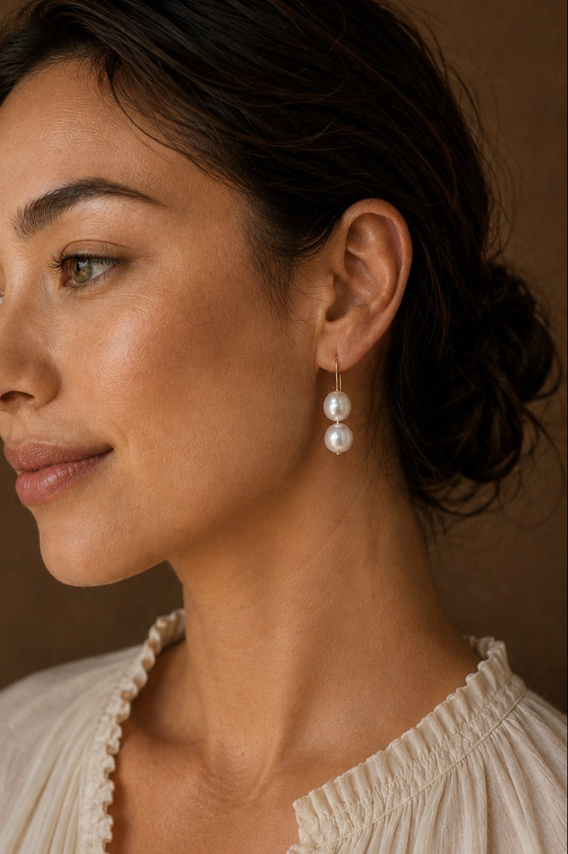 A woman in profile wearing a pair of double pearl drop earrings, set against a warm brown background.