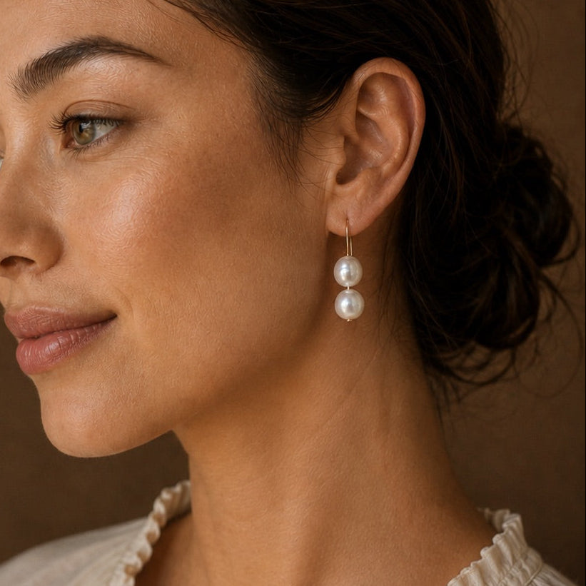 A woman in profile wearing a pair of double pearl drop earrings, set against a warm brown background.