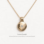 A gold pendant necklace with a flat, disc-shaped pendant featuring a small, centered, round-cut diamond. The necklace is displayed on a clean white background with descriptive text below.