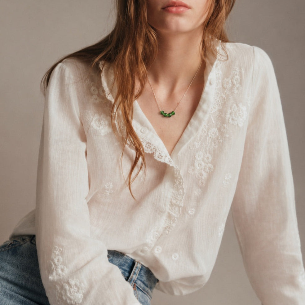 A woman wearing a white long-sleeve blouse with delicate lace embroidery and ruffled cuffs, paired with blue jeans, posed against a neutral beige studio background.
