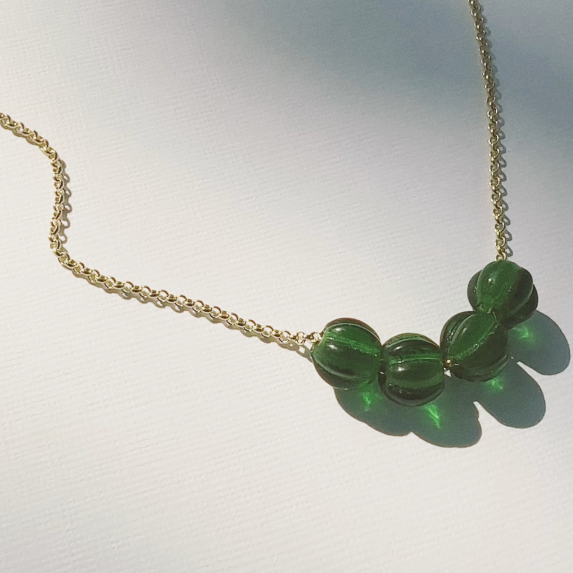 A gold chain necklace featuring three faceted, translucent green glass beads clustered on one end, displayed on a white textured surface with soft shadows.
