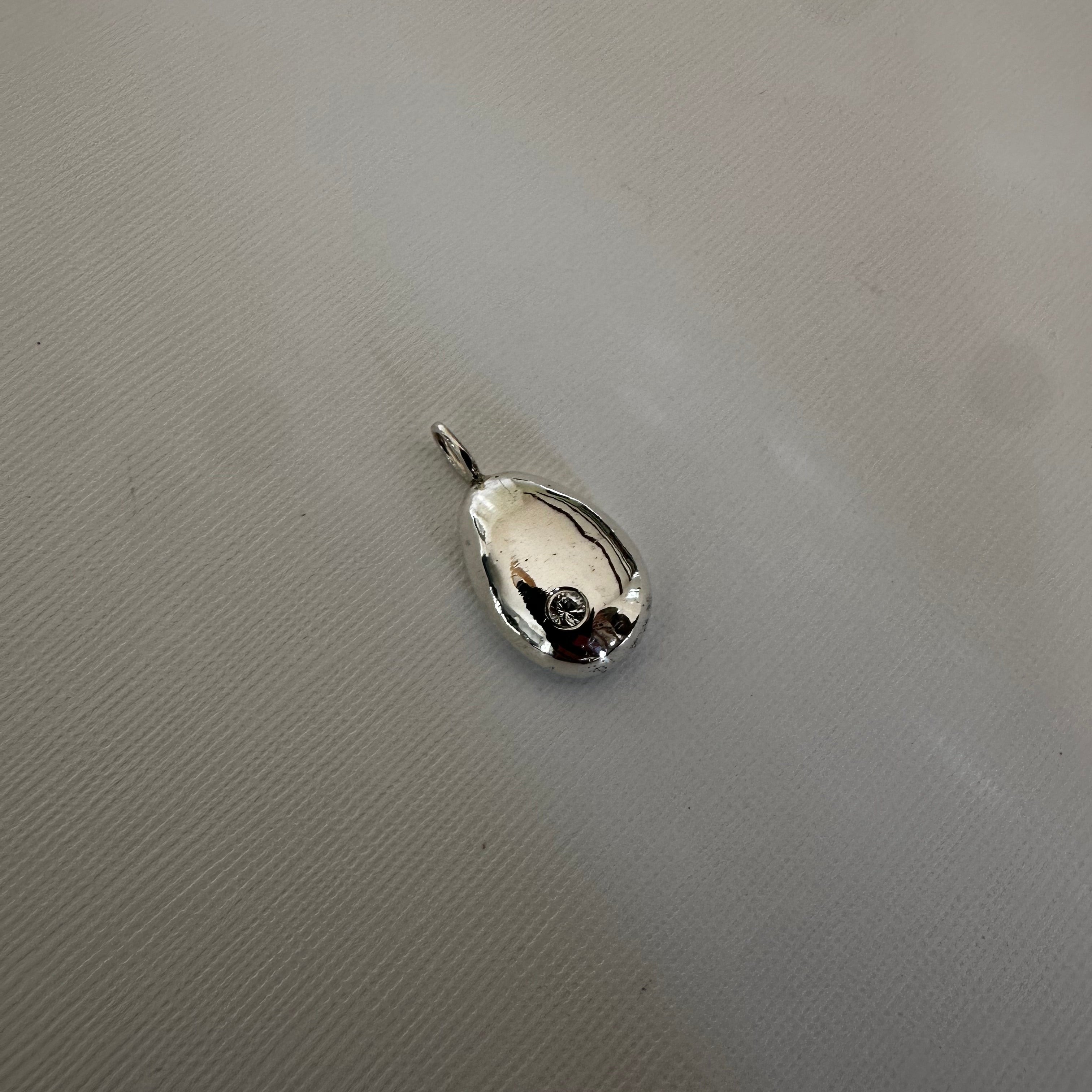 A small polished silver teardrop-shaped pendant or charm with a short loop at the top, placed on a textured light gray surface.