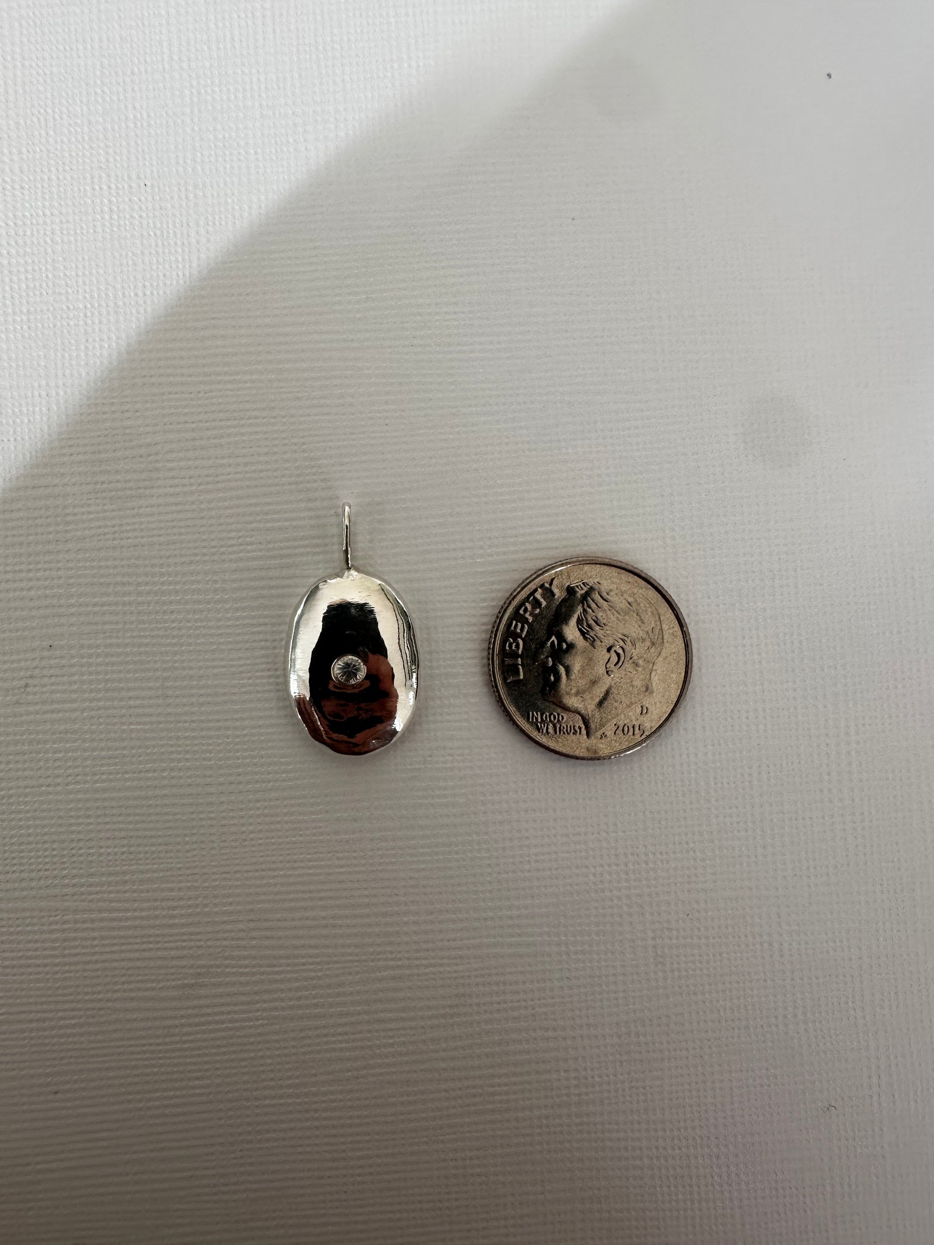 A small polished silver teardrop-shaped pendant or charm with a loop at the top, shown next to a U.S.