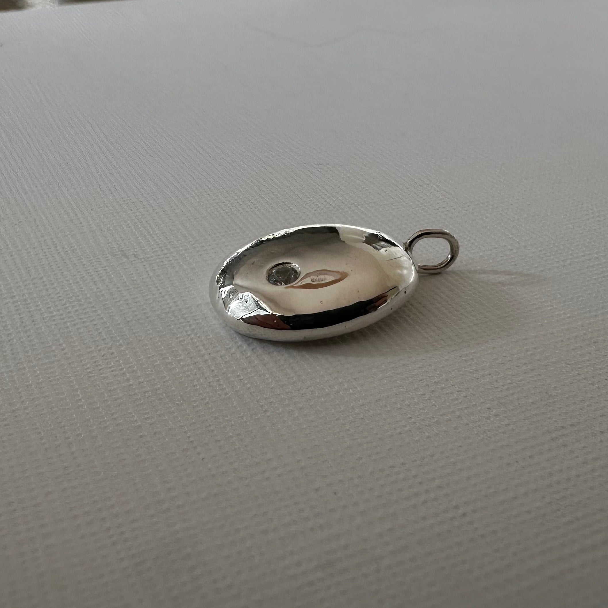 A polished silver oval pendant with a small hanging loop, resting on a textured white surface.