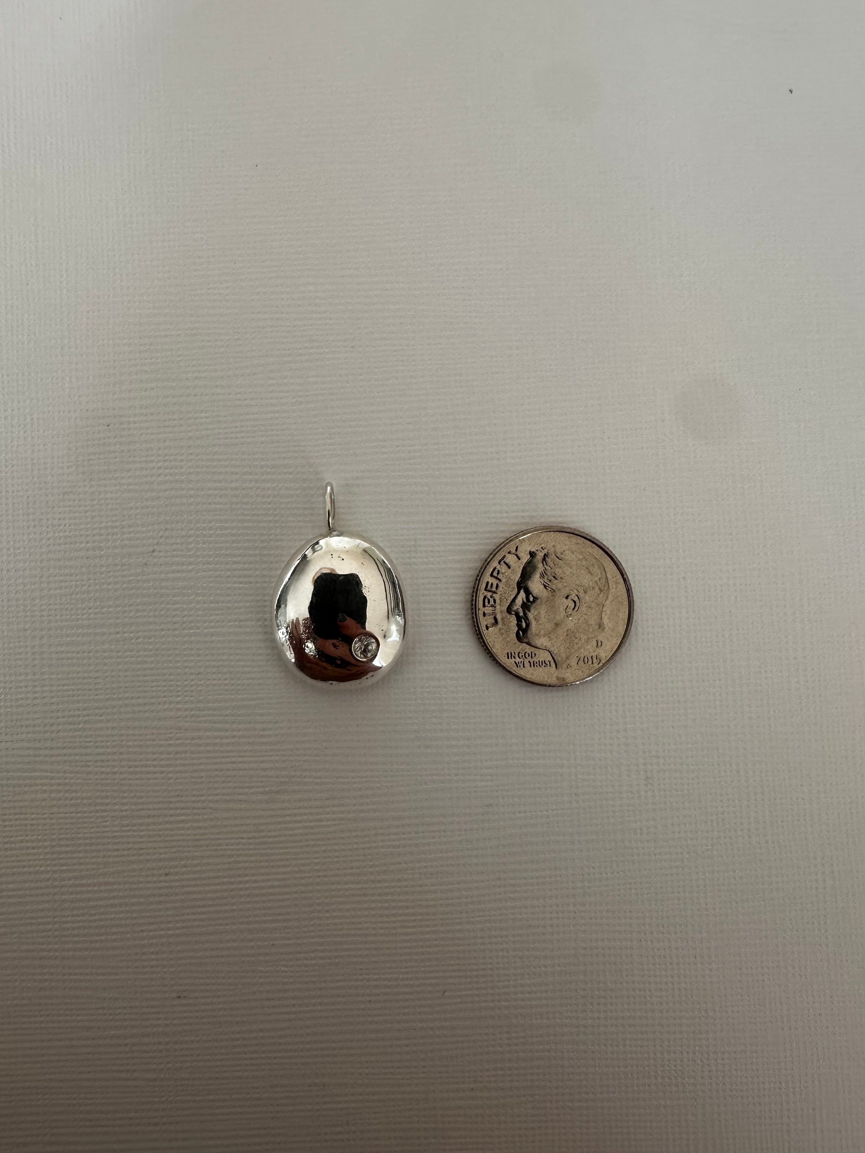 A small silver teardrop-shaped pendant with a reflective surface, shown next to a US quarter coin for scale on a light gray textured background.