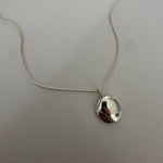 A silver necklace with a small, irregularly shaped polished pendant on a delicate chain, laid flat on a textured light gray surface.