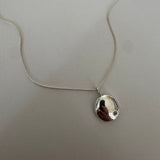 A silver necklace with a small, irregularly shaped polished pendant on a delicate chain, laid flat on a textured light gray surface.