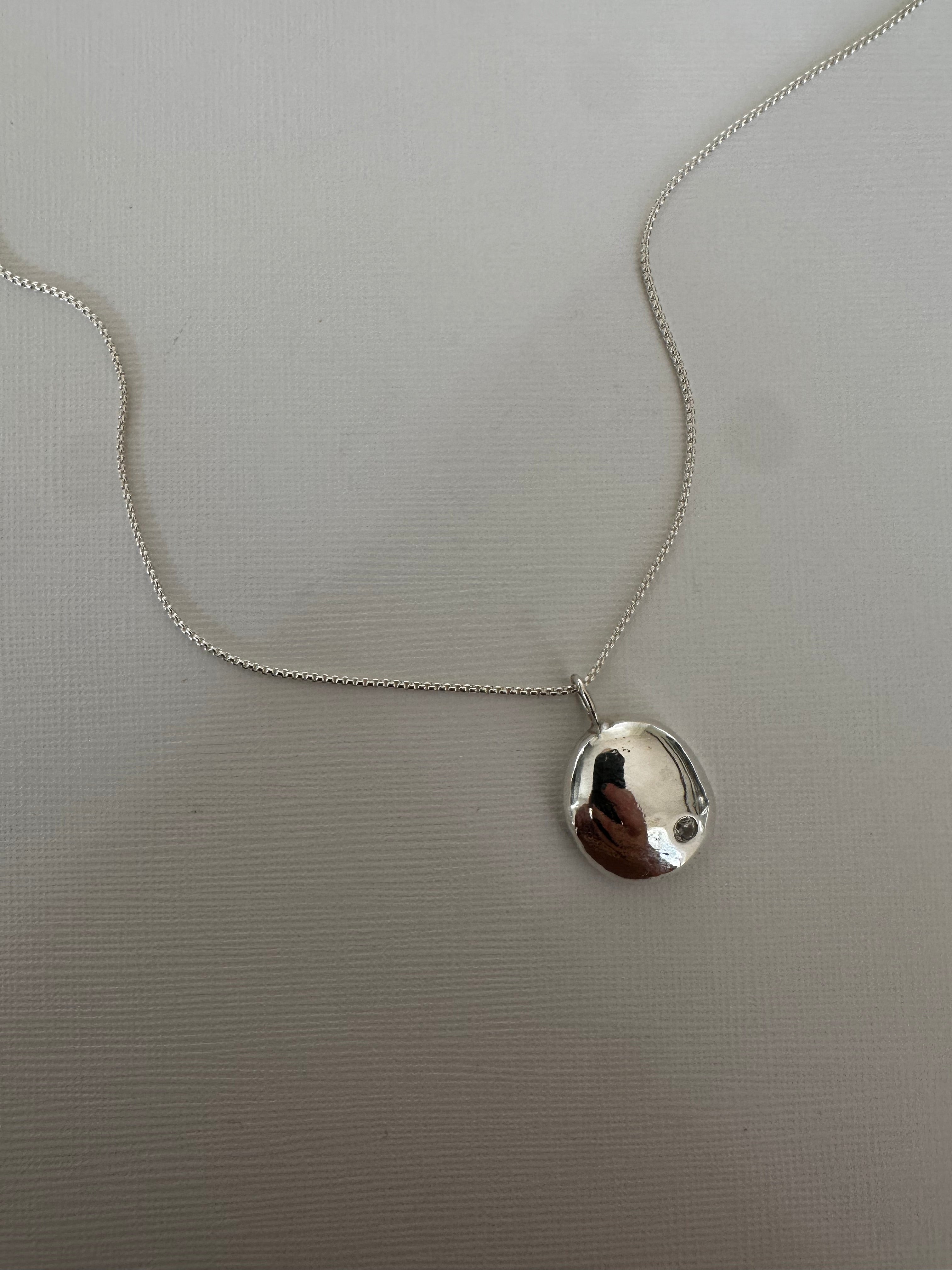 A silver necklace with a small, irregularly shaped polished pendant on a delicate chain, laid flat on a textured light gray surface.