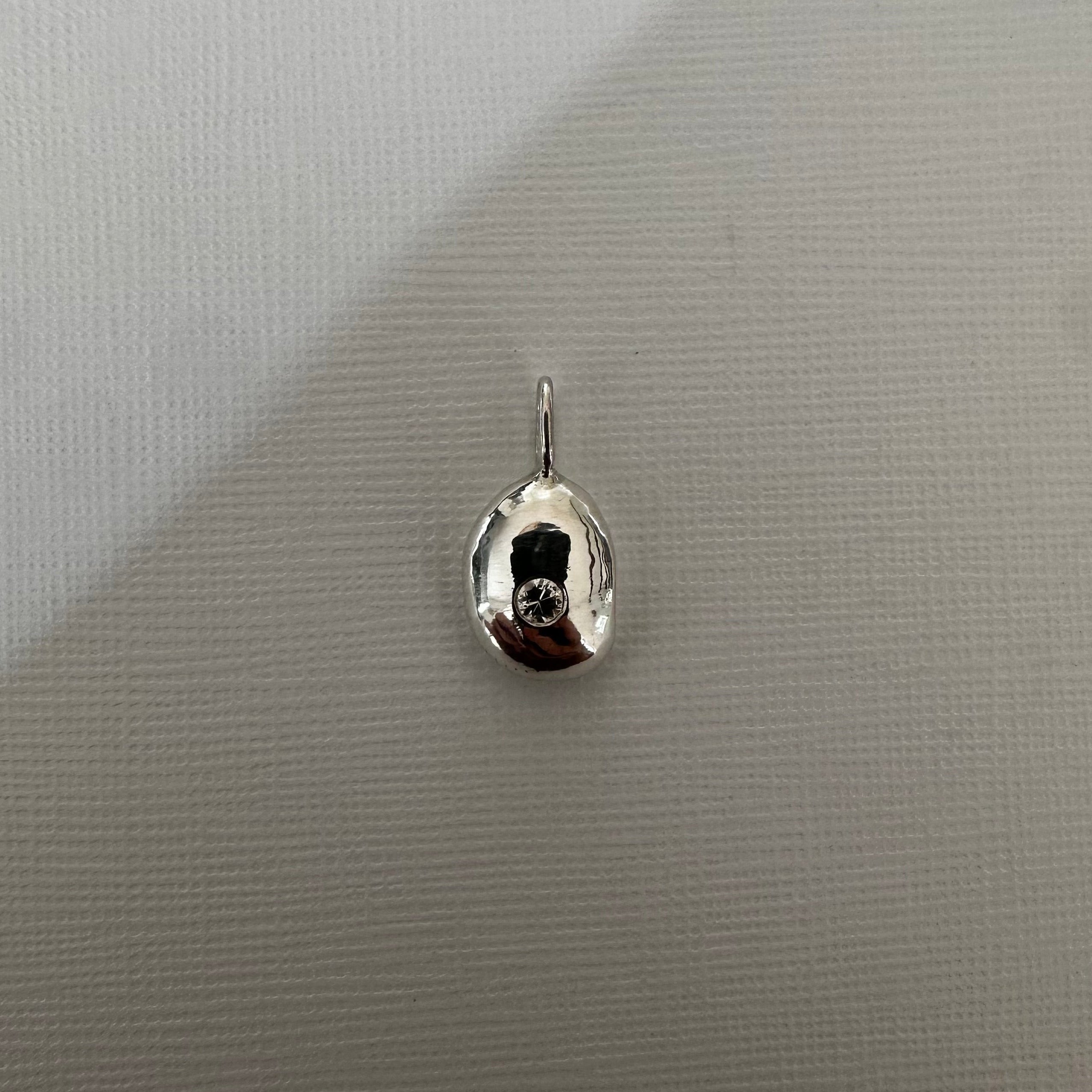 A small polished silver teardrop-shaped pendant or charm with a reflective finish and a tiny loop at the top, shown on a light gray textured background.