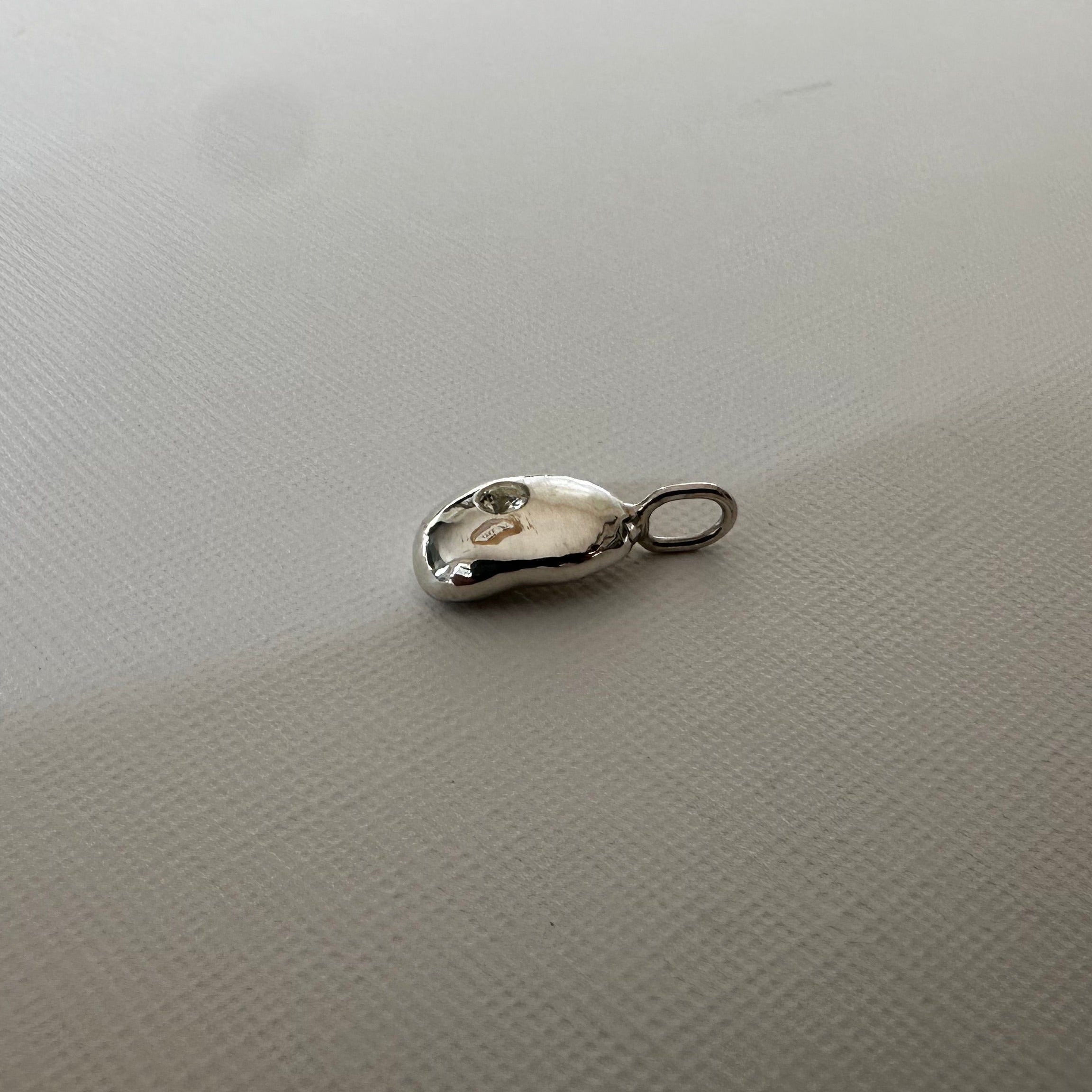A small silver metal charm or pendant with a loop at one end, resting on a textured off-white surface.