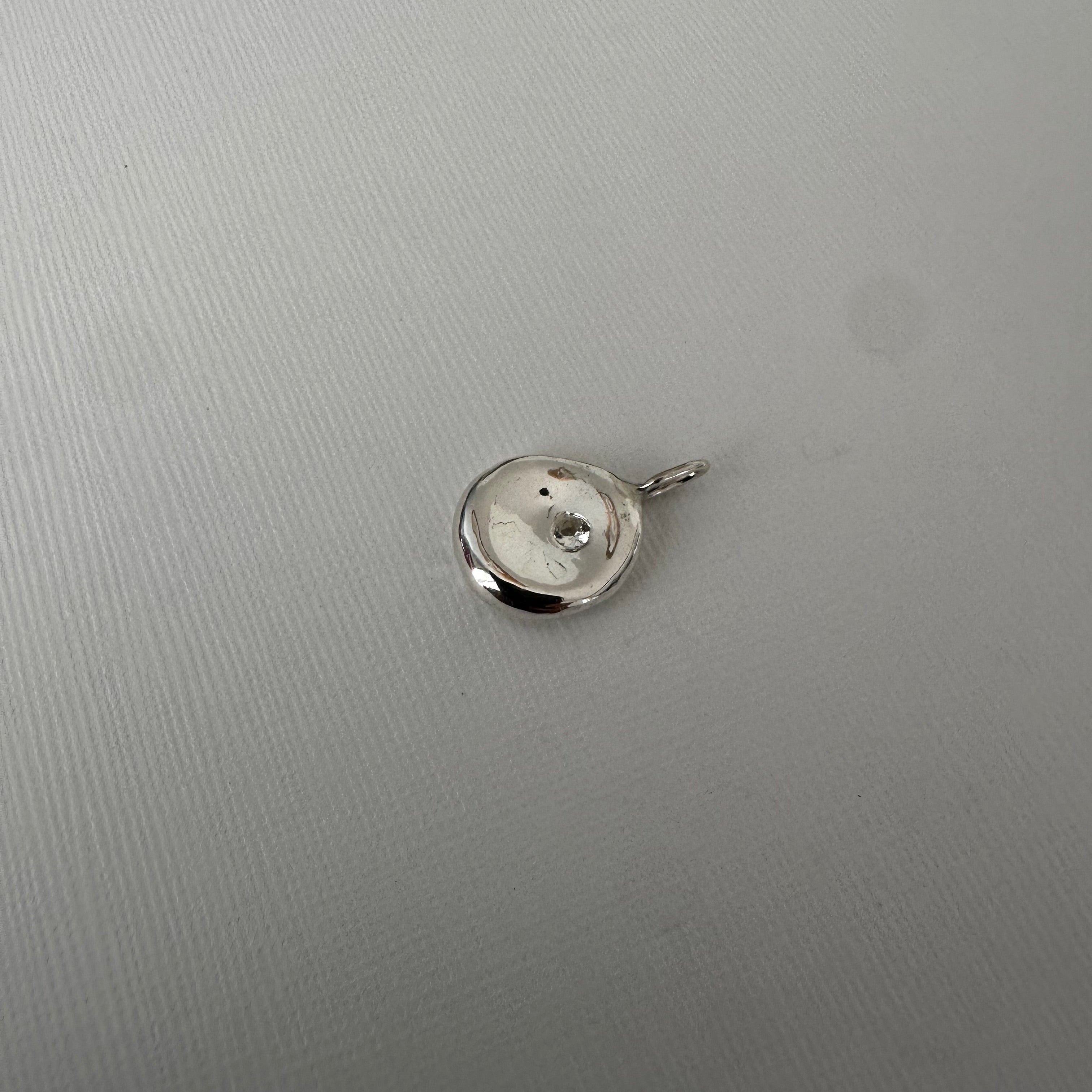 A small polished silver pendant or charm with a circular disc design and a tiny central dot, shown on a light gray textured background.