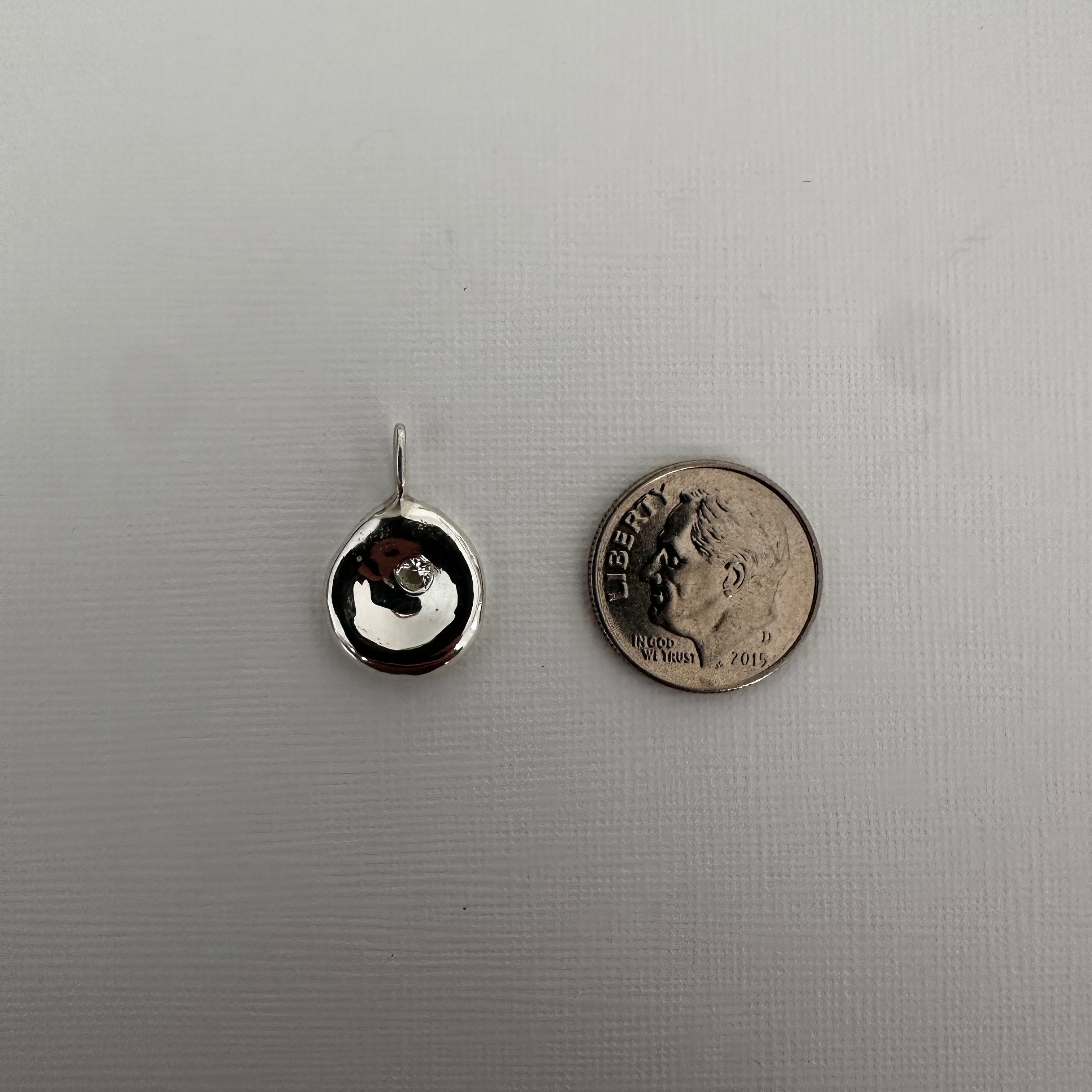 A small silver stud earring with a circular face and a thin post, shown on a light gray textured background with a US quarter coin above it for scale.