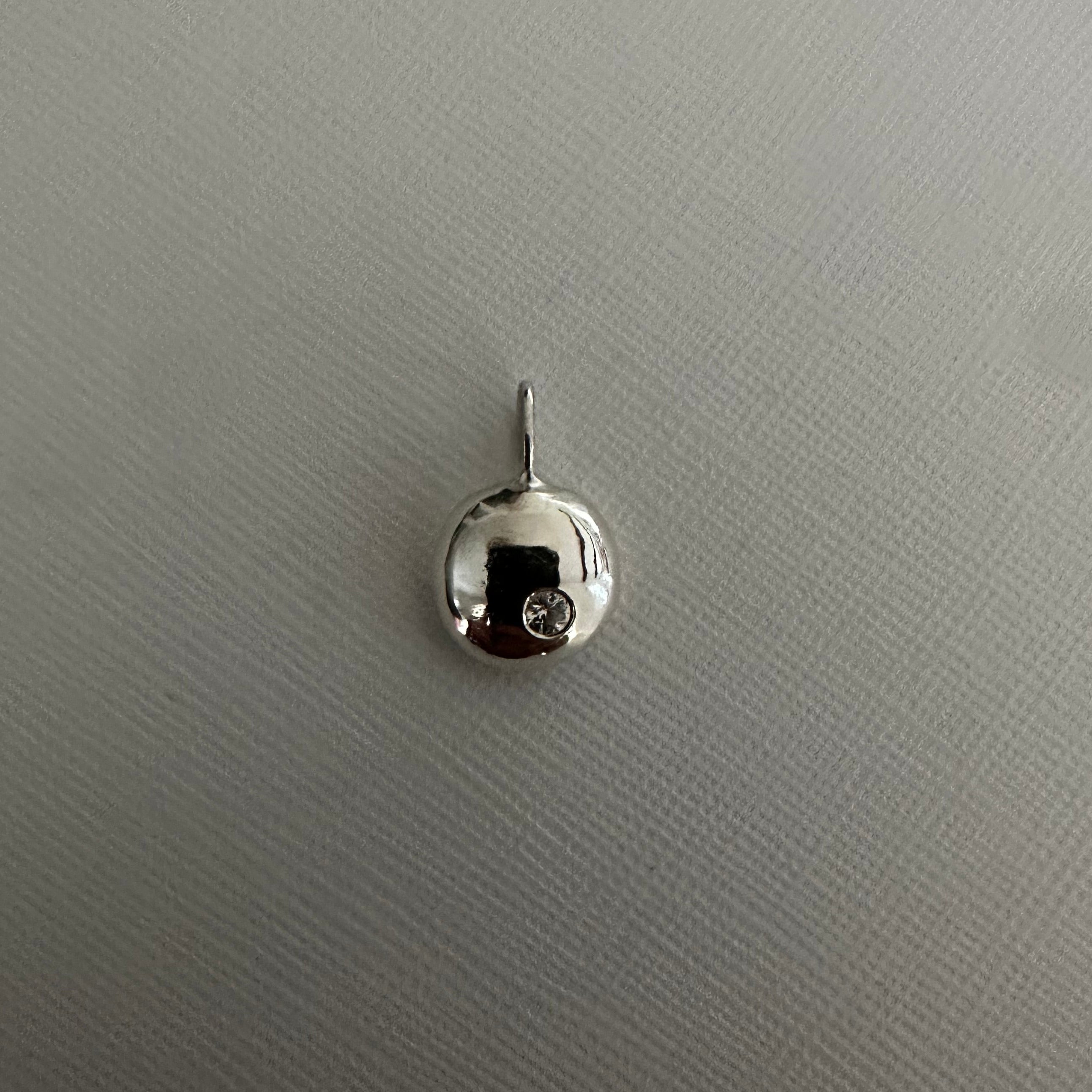 A small polished silver pendant or charm with a reflective dome-like surface and a short loop at the top, shown on a light gray textured background.