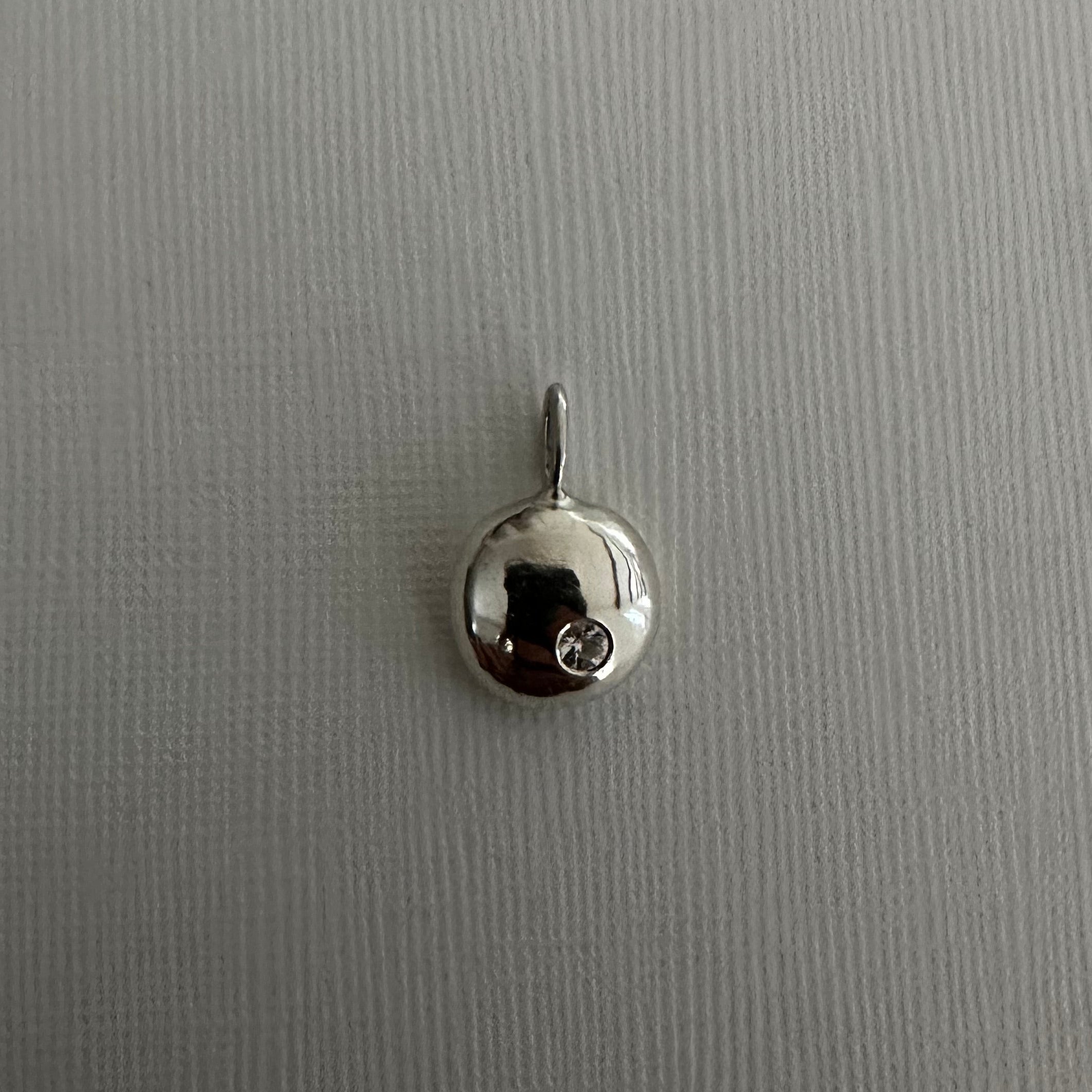 A small silver pendant charm with a polished dome and a tiny clear gemstone, shown on a textured gray background.