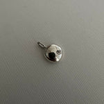 A small silver charm pendant with a flat circular disc design and a tiny clear stone, shown on a textured light gray background.