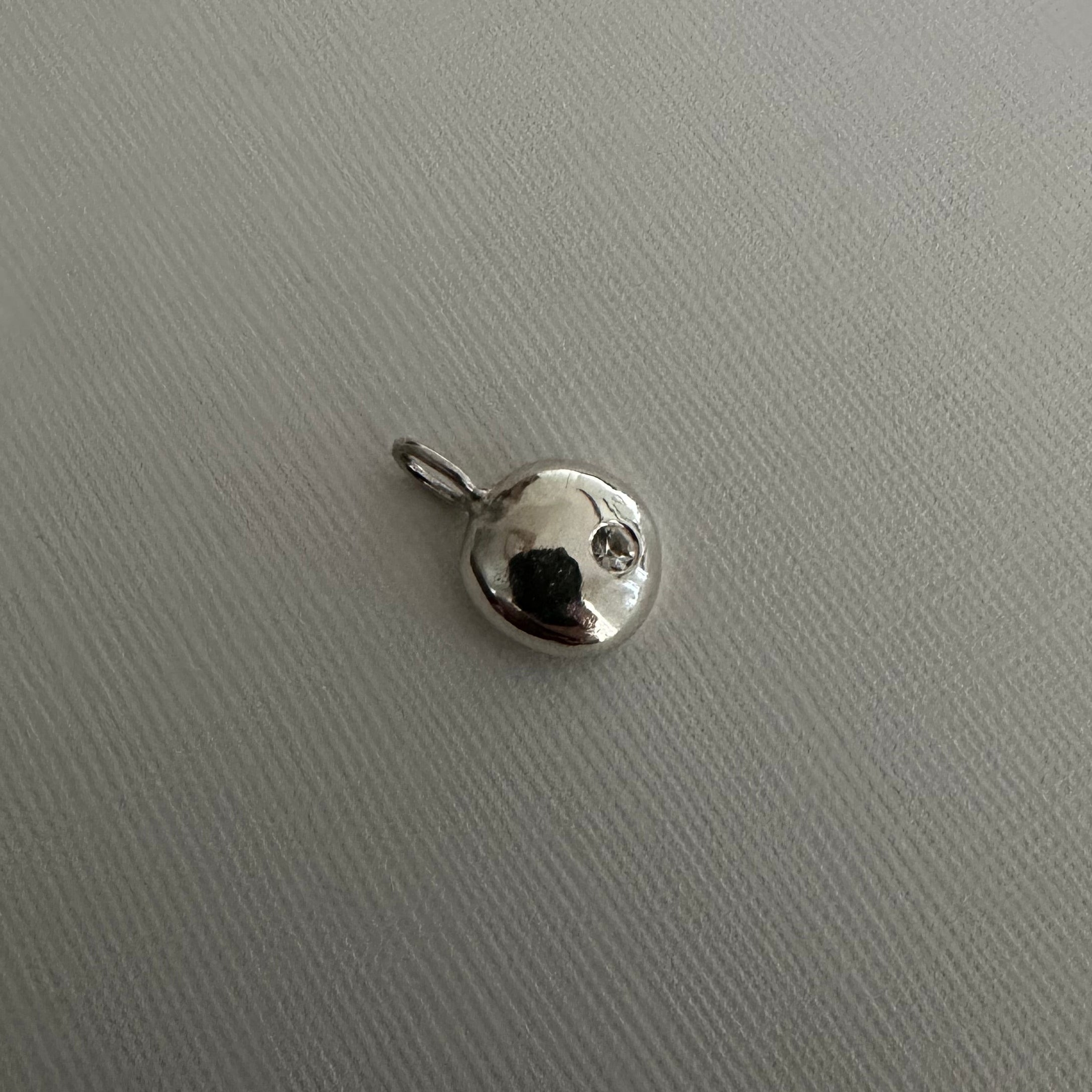 A small silver charm pendant with a flat circular disc design and a tiny clear stone, shown on a textured light gray background.