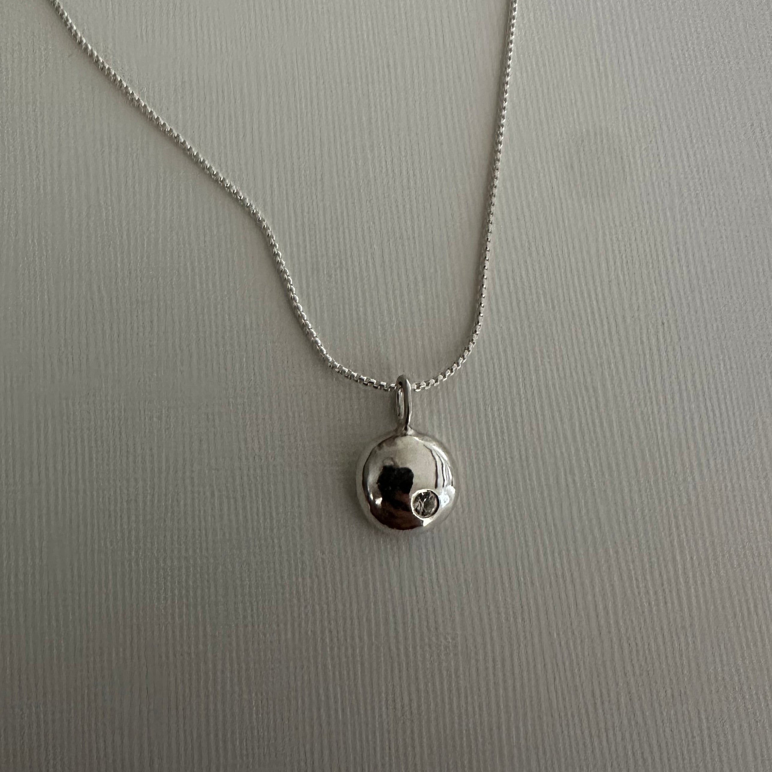 A silver beaded necklace with a small round silver bead pendant on a textured light gray background.