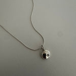 A silver chain necklace with a small, polished silver disc pendant featuring a black center, displayed on a textured light gray surface.