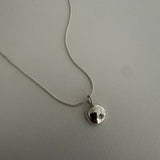 A silver chain necklace with a small, polished silver disc pendant featuring a black center, displayed on a textured light gray surface.