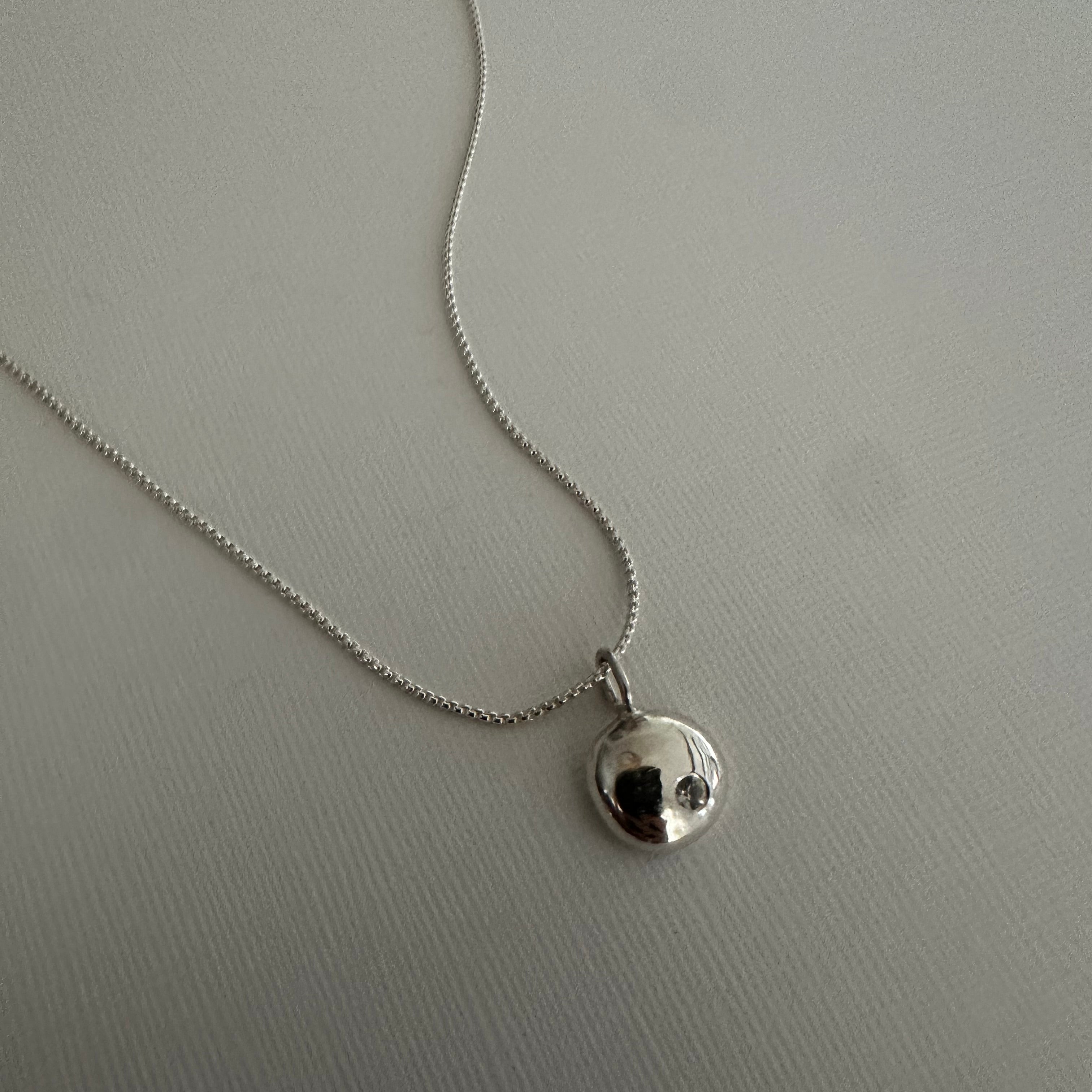 A silver chain necklace with a small, polished silver disc pendant featuring a black center, displayed on a textured light gray surface.