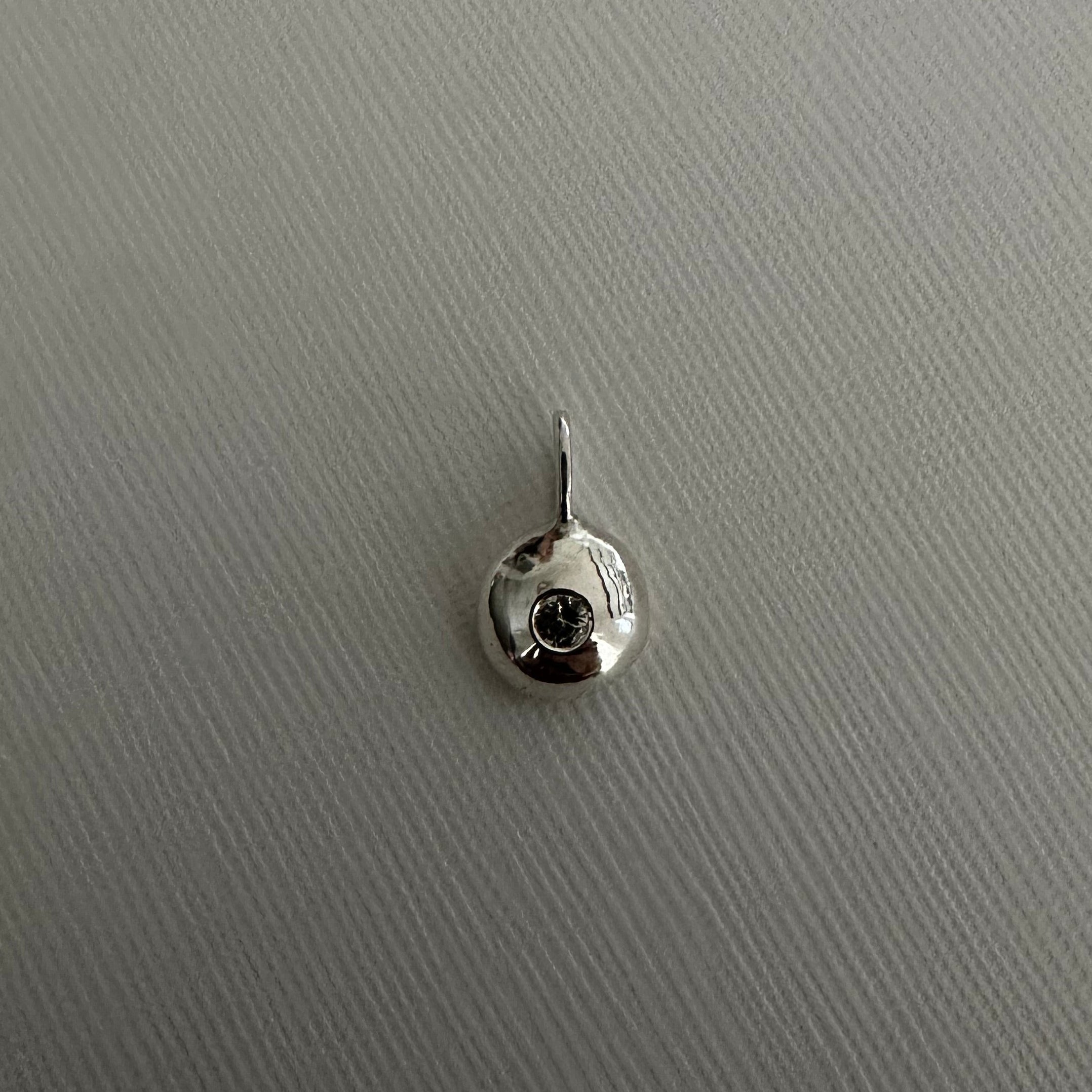 A small silver circular stud earring with a central clear gemstone and a top prong setting, shown on a textured light gray background.
