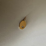 A small oval gold pendant with a tiny clear gemstone set in the center, resting on a textured off-white fabric background.