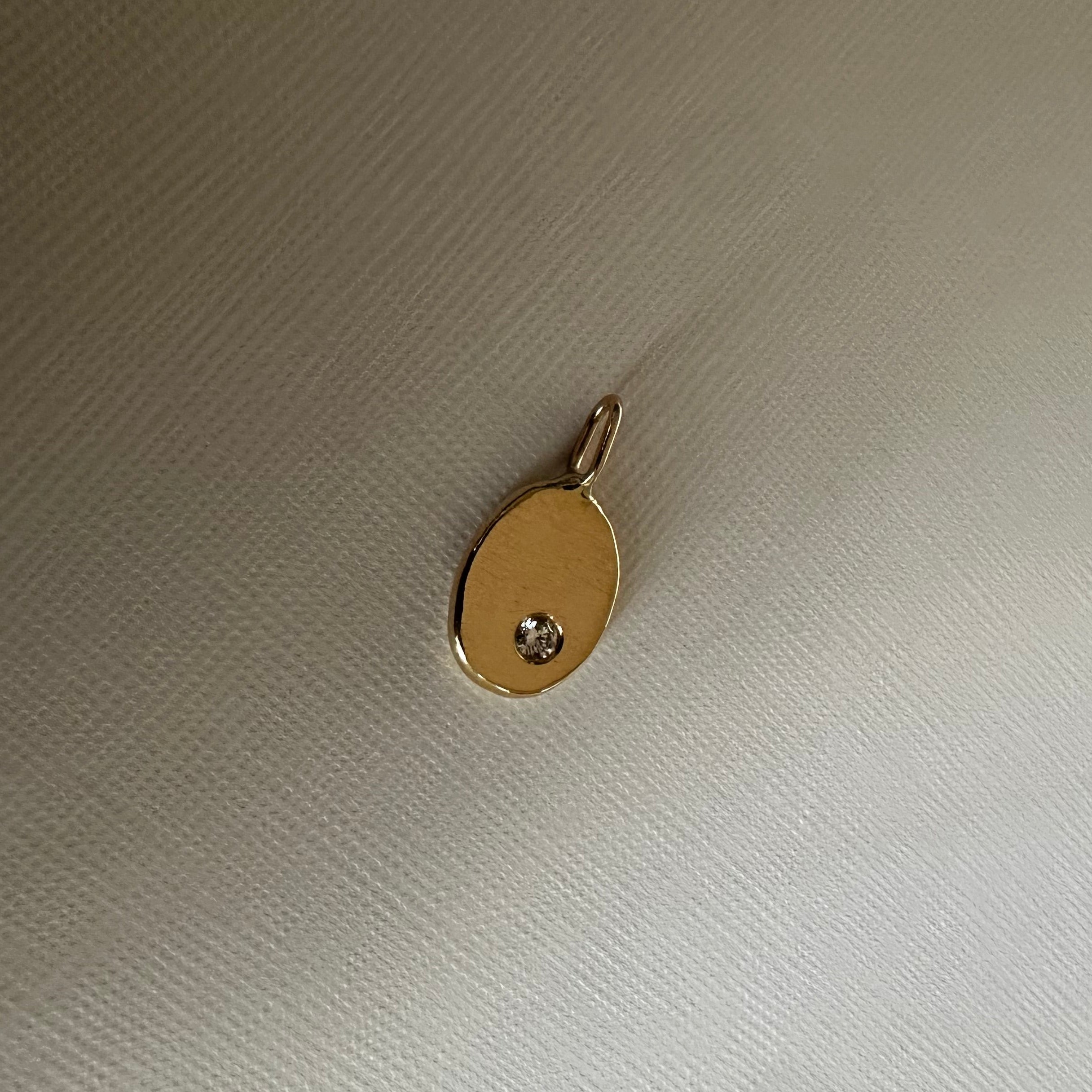 A small oval gold pendant with a tiny clear gemstone set in the center, resting on a textured off-white fabric background.