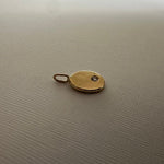 A small oval gold pendant or charm with a loop at the top and a tiny dark stone inset, placed on a textured light beige surface.