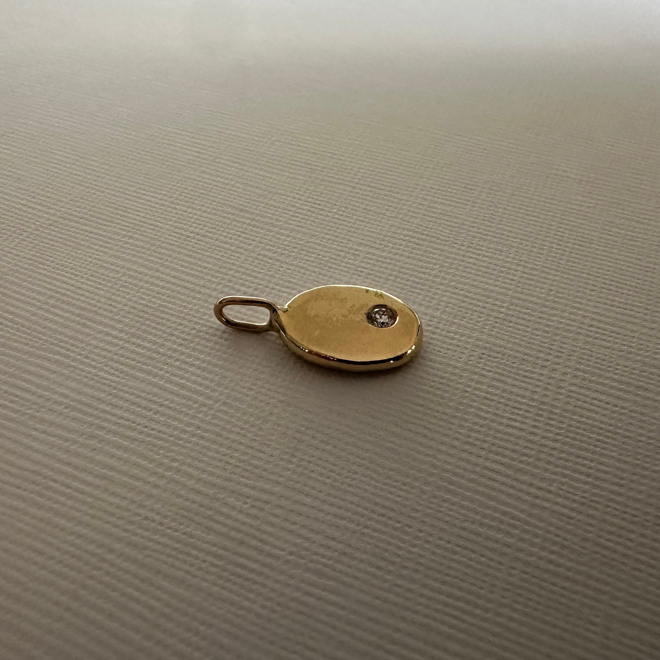 A small oval gold pendant or charm with a loop at the top and a tiny dark stone inset, placed on a textured light beige surface.