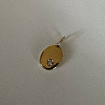 A small gold oval pendant with a tiny clear gemstone set in one corner, resting on a textured off-white fabric background.