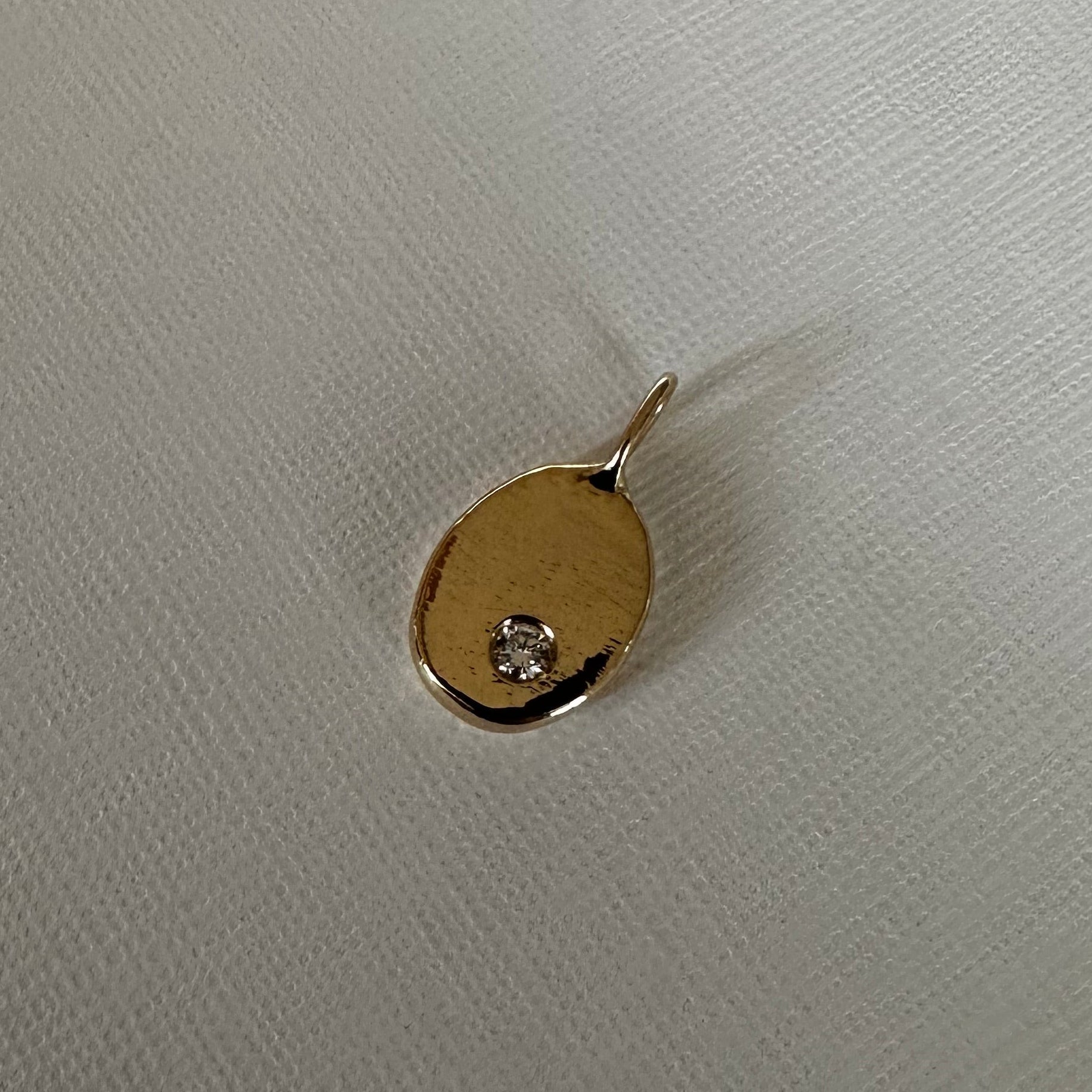 A small gold oval pendant with a tiny clear gemstone set in one corner, resting on a textured off-white fabric background.