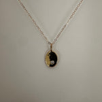 A delicate gold chain necklace with an oval pendant featuring a black and gold yin-yang symbol and a small clear gemstone, displayed against a light gray background.
