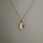 A delicate cable gold chain necklace with a small, hammered oval pendant, displayed against a neutral gray textured background.