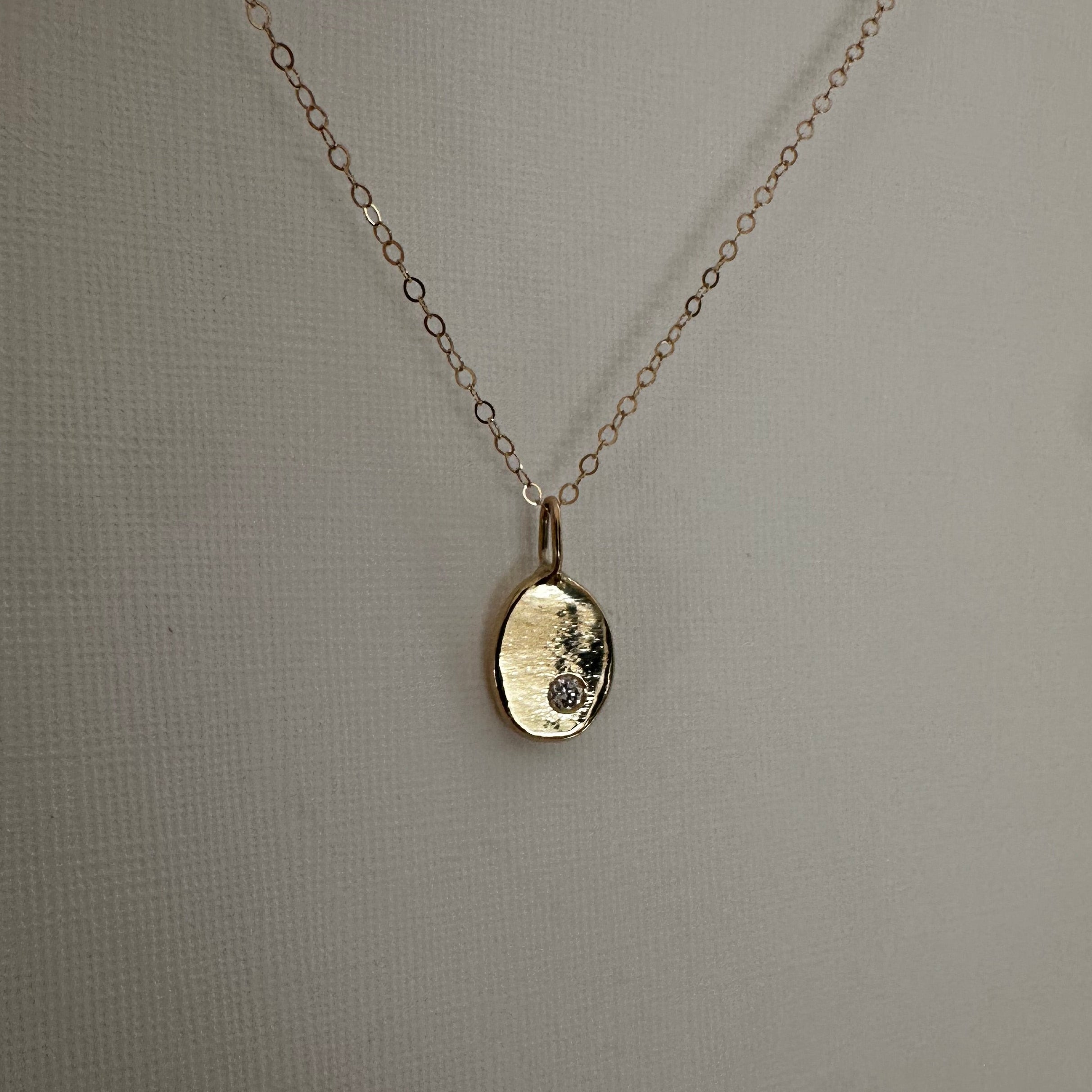 A delicate cable gold chain necklace with a small, hammered oval pendant, displayed against a neutral gray textured background.