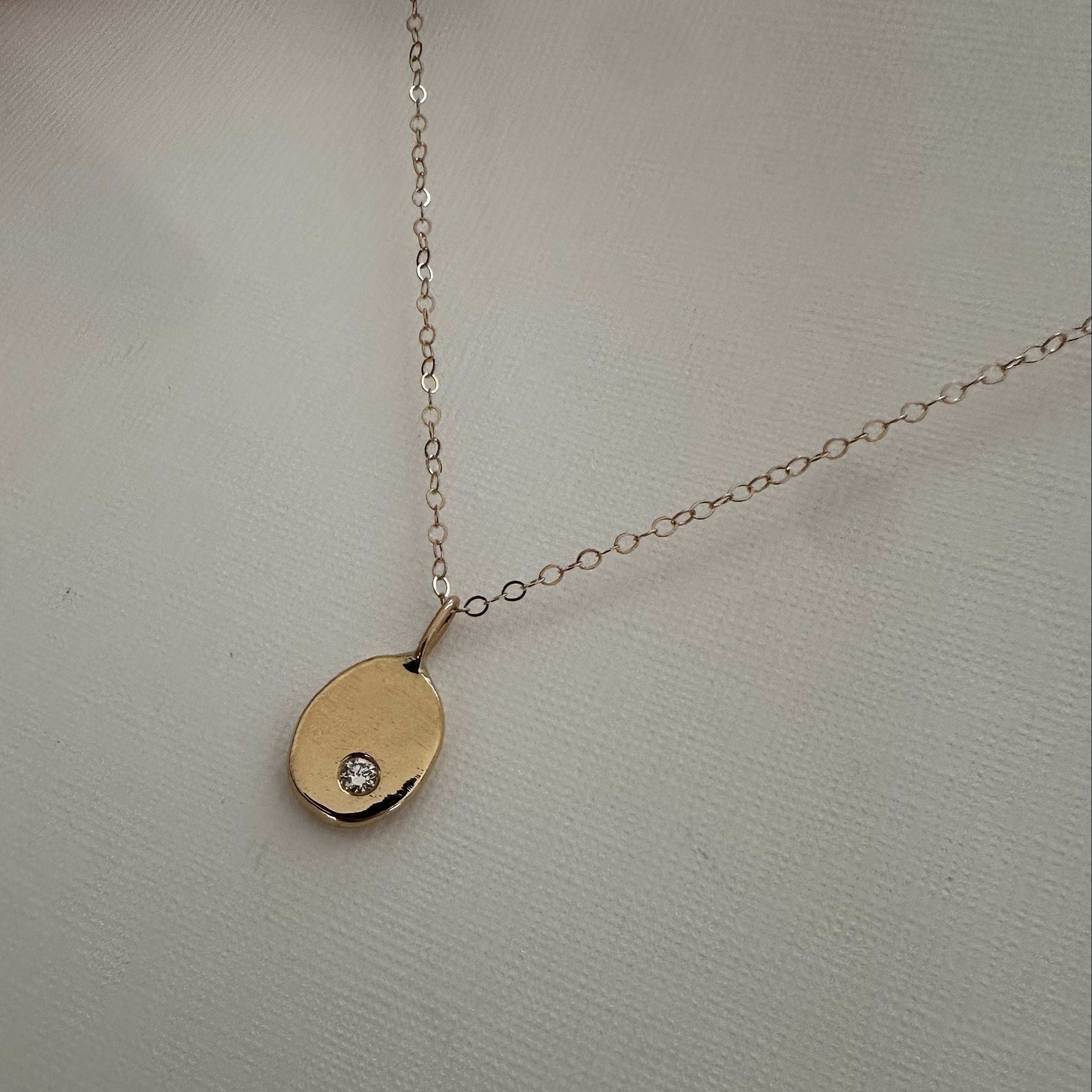 A delicate gold chain necklace with an oval gold pendant featuring a small clear gemstone, displayed on a textured off-white surface.