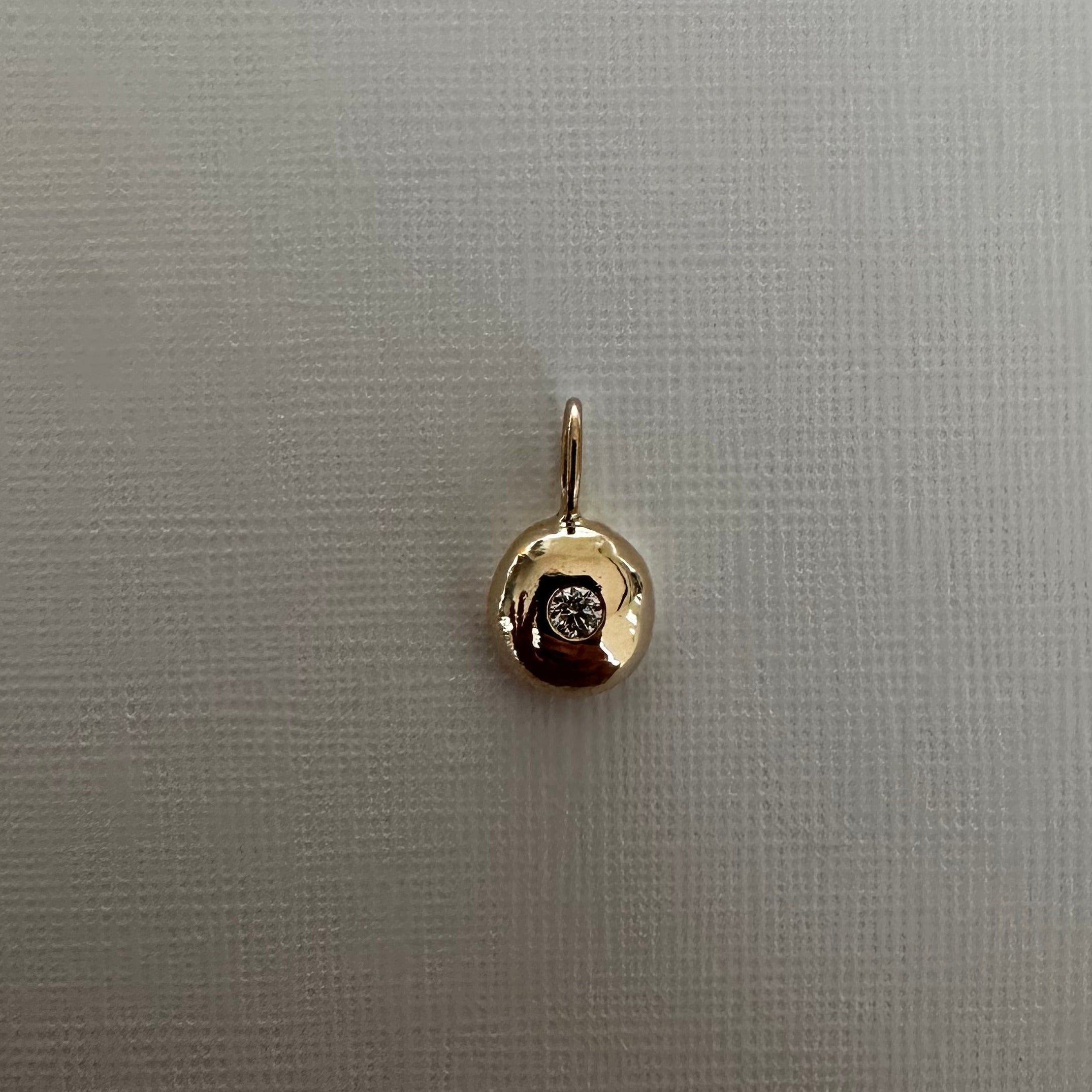 A small gold pendant charm with a circular textured design and a central dark stone, displayed on a light gray textured fabric background.