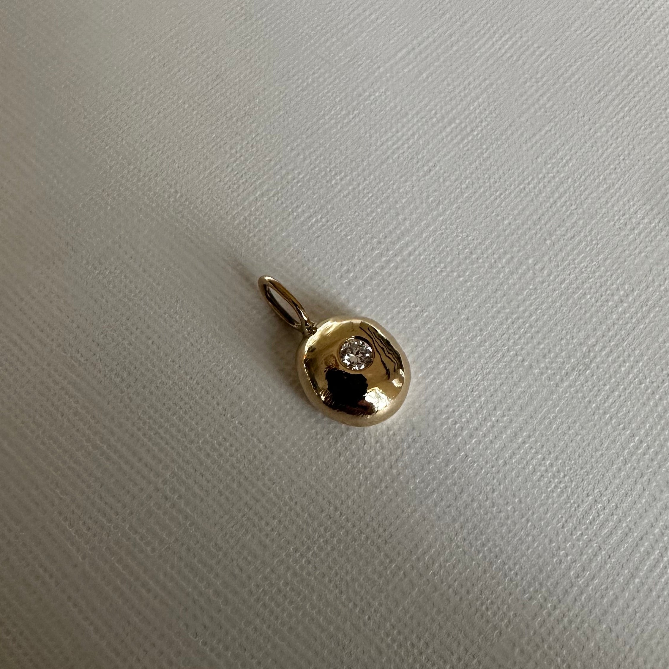 A small gold pendant with a tiny clear gemstone, resting on a textured off-white fabric surface.