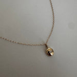 A delicate  yellow gold chain necklace with a small, textured circular pendant featuring a tiny clear gemstone, displayed on a light beige textured fabric background.